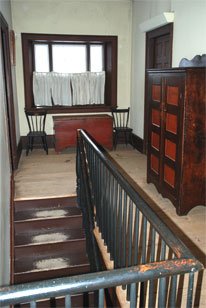 The Inn's second floor hallway, has a window at the top of the steps. The worn stairs leading up to the second floor are visible beyond the banister. Their stain has worn through to the bare wood in the middle of each step. A large armoire sits in the hallway, parallel to the stairs, on the other side of the banister.