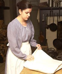 A Montgomery's Inn servant in period dress, folding fabric on a table in the kitchen.